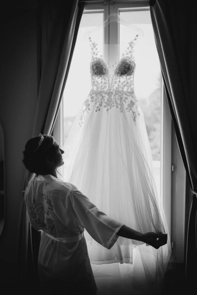 La mariée touche sa robe suspendue à la fenêtre - Photographe mariage Yvelines